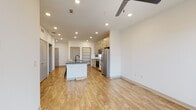 This single-floor space showcases a contemporary open-concept design with a cohesive, neutral color palette and light wood-look flooring that runs consistently throughout. The kitchen serves as the centerpiece of the main living area, equipped with light wood cabinetry extending to the ceiling, a white subway tile backsplash, and a full suite of stainless steel appliances including a refrigerator, range, and over-the-range microwave. A center island with a dark quartz countertop and an undermount sink with a matte black faucet provides additional workspace and casual seating. Open shelving built into a dedicated nook offers practical storage and display space adjacent to the kitchen. Recessed lighting throughout the main living area is supplemented by a ceiling fan, ensuring both functional illumination and air circulation. The bedroom is a well-proportioned, bright room featuring two windows with blinds that allow natural light, paired with a ceiling fan overhead. The bathroom features wood-look flooring, a modern vanity with a dark countertop, textured cabinetry, and a tub-shower combination finished with white subway tile and matte black fixtures. The overall ambiance is clean, modern, and functional.