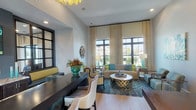 This thoughtfully designed single-floor common area blends contemporary elegance with warm, inviting touches throughout its distinct zones. The main lounge area makes a strong visual statement with bold teal and gold accents, a striking geometric chandelier, and floor-to-ceiling patterned drapery framing large windows that flood the space with natural light and offer outdoor views. A dark espresso desk with cream upholstered chairs anchors a functional workspace within this vibrant setting, complemented by a decorative sunburst wall mirror and lush floral arrangements.

A separate seating area provides a more relaxed atmosphere, centered around a dramatic floor-to-ceiling stacked stone fireplace with a rustic wood mantel. Comfortable upholstered chairs are arranged to encourage conversation, while French doors with divided-light panes open directly to an outdoor space, enhancing the flow between interior and exterior areas.

An additional lounge section features a cream tufted sofa accented with soft teal and gold pillows, accompanied by wingback chairs and small accent tables. A wall-mounted flat-screen television and tasteful artwork complete this area. Throughout, tall ceilings, recessed lighting, multi-arm chandeliers, and neutral wall tones create a cohesive, airy ambiance.