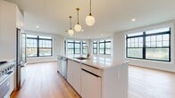 Located at 695 Merrick Ave in Westbury, NY, this thoughtfully designed unit showcases a seamless blend of modern aesthetics and high-end finishes across a single, well-appointed floor. Wide-plank natural hardwood flooring flows throughout every room, lending warmth and continuity to the space.

The open-concept kitchen serves as a central showpiece, featuring a large center island with a white quartz countertop, an undermount sink with a brushed gold faucet, and sleek flat-front white cabinetry extending to the ceiling for ample storage. Stainless steel appliances, including a refrigerator and microwave, complement the contemporary design. A trio of globe pendant lights with brass fixtures hang above the island, adding an elegant touch. Black-framed multi-pane windows surround the adjacent living area, filling the space with abundant natural light and offering open views of the surroundings.

The hallways connect generously sized rooms, with white interior doors accented by gold hardware throughout. The bathroom is finished with large-format marble-look porcelain tile flooring, a wood-framed vanity with a stone countertop and gold fixtures, and a frameless glass walk-in shower clad in textured subway tile with a built-in bench and brushed gold fixtures. The overall ambiance is clean, bright, and contemporary.