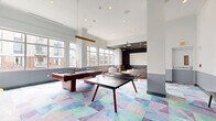 Located at 111 Towne St in Stamford, Connecticut, this single-floor amenity lounge is a thoughtfully designed recreational space that seamlessly blends entertainment, comfort, and contemporary style. The space is defined by soaring ceilings and large windows that line the walls, flooding the interior with natural light and offering views of the surrounding urban streetscape.

The main game area serves as the centerpiece, featuring both a pool table and a ping-pong table positioned atop a striking geometric-patterned carpet in soft hues of lavender, teal, and sage. This colorful flooring element anchors the space and adds a lively, modern character. Comfortable lounge seating is arranged nearby, providing a relaxed area for socializing, while a wall-mounted flat-screen TV adds to the entertainment options.

A dedicated screening room is tucked within the space, distinguished by its deep navy blue walls and a dark ceiling with recessed lighting that creates a true cinematic atmosphere. The room is furnished with plush sofas, floor cushions, and a round ottoman centered around a large projection screen, making it ideal for group viewings.

Decorative gold wall-mounted hooks and sleek dark benches add refined touches throughout, complementing the two-tone gray and white wall treatment. Light hardwood flooring in the screening area contrasts beautifully with the patterned carpet of the game zone, tying the entire space together with a polished, cohesive aesthetic.