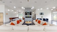 Located at 888 Horizon Boulevard in Athens, Georgia, this single-floor community lounge is a generously sized, open-plan gathering space designed with a contemporary and energetic aesthetic. Polished concrete floors run throughout, reflecting natural light that pours in through large floor-to-ceiling windows offering views of the outdoor pool area. Soaring ceilings are adorned with an array of globe pendant lights, creating a striking visual canopy that defines the space's modern character. A bold, black-and-white geometric accent wall serves as the room's focal point, housing a sleek linear fireplace and a mounted flat-screen television. The main seating area features oversized white sectional sofas accented with vibrant orange throw pillows, centered around a round coffee table. Additional seating includes plush navy blue and orange velvet chairs, providing diverse lounge options. A dedicated dining nook with modern chairs and a bistro-style table sits near the windows. A games area with a pool table adds a recreational element, while a retro-inspired "Good Vibes" wall mural injects personality and fun. The white walls and bright, open layout create an inviting, social atmosphere ideal for gathering and relaxation.
