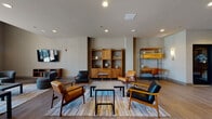 Located at 1188 Main St in Bridgeport, Connecticut, this thoughtfully designed community space blends contemporary aesthetics with warm, functional interiors. The entrance corridor immediately impresses with large-format tile flooring and a striking skylight that floods the passage with natural light, creating an open and welcoming atmosphere as you move through the space.

The main lounge area is generously proportioned, featuring warm hardwood-style flooring, recessed lighting, and mid-century modern furnishings including leather and wood-frame chairs arranged around low coffee tables. Open wood shelving units provide both display space and architectural character, while wall-mounted televisions offer entertainment throughout. Colorful area rugs anchor the seating zones and add vibrancy to the neutral palette.

A dedicated lounge foyer features a textured metallic accent wall paired with a round mirror and a handsome wood console table, creating an elegant, curated vignette. Nearby, a communal bar and café area offers an extended granite-topped island with leather bar seating, pendant lighting, and a built-in coffee station — ideal for casual gatherings or individual use. Textured stone-effect wall treatments lend an upscale, modern character to this zone.

Additional dining seating with sconce-lit walls and a welcoming lobby with a stone-accented elevator bay round out this well-appointed community floor.