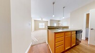 This single-floor residence offers a thoughtfully designed layout that maximizes every square foot across its living room, kitchen, bedroom, bathroom, and laundry areas. The open-concept kitchen and living area form the heart of the home, featuring warm honey-toned cabinetry, a double-basin sink set into a peninsula with dark laminate countertops, and a full suite of black appliances including a refrigerator, range, and over-the-range microwave. Rich hardwood-style flooring runs through the kitchen and dining area, while plush neutral carpeting flows throughout the living room and bedroom, adding warmth and comfort underfoot. The living room is generously proportioned, bathed in natural light from multiple windows, and accented by a ceiling fan overhead. A dedicated dining area features an elegant chandelier and additional window views. The spacious primary bedroom includes a walk-in closet with built-in shelving and benefits from natural light through its own window. The bathroom is finished with tile flooring, a full soaking tub with tiled surround, and a rich wood-toned vanity. Warm beige and neutral tones are carried throughout the space, creating a cohesive and inviting ambiance.