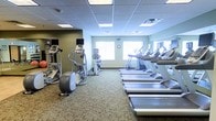 This fitness center offers a dedicated space for cardiovascular exercise, thoughtfully laid out to maximize usability and comfort. A row of treadmills lines one side of the room, providing ample options for users during peak hours, while a collection of elliptical trainers and a stationary bike occupies the opposite side, catering to a range of low-impact workout preferences. Each cardio machine is equipped with an individual monitor, allowing users to track performance metrics or enjoy entertainment during their sessions. The room is finished with a neutral, patterned carpet that adds a degree of comfort underfoot. Soft green walls contribute to a calm, focused atmosphere. Natural light enters through several windows along the far wall, brightening the interior and creating an open feel. Recessed panel lighting in the drop ceiling ensures consistent illumination throughout the space. A wall clock is centrally positioned, keeping exercisers mindful of their time. The overall layout is clean and organized, offering a functional and accessible environment well-suited for daily fitness routines.