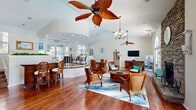 Located at 1088 West Granada Boulevard in Ormond Beach, Florida, this single-floor community clubhouse offers a generously sized, open-concept gathering space with a warm coastal aesthetic. The main lounge area is anchored by a dramatic floor-to-ceiling stacked stone fireplace, complemented by soaring vaulted ceilings, rich hardwood flooring, and abundant natural light streaming through arched French doors and large windows. The coastal-inspired furnishings — including wicker and rattan chairs with teal accent pillows — create a relaxed, inviting atmosphere throughout.

A curved, raised bar area with a coffered ceiling and recessed lighting serves as a central architectural focal point, seamlessly separating the lounge from the dining and gathering zones. Multiple ceiling fans, including a distinctive tropical palm-blade fan, enhance the breezy Florida ambiance. A built-in bar counter with elegant carved wood barstools adds a refined social space for residents.

The French doors open to outdoor views of a pool area, reinforcing the indoor-outdoor connection. A separate, carpeted office area features a built-in granite-topped desk with cabinetry, providing a functional workspace. The space is further enhanced by a wall-mounted television, ample seating arrangements, and lush indoor greenery, making it a versatile and comfortable hub for community life.