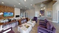 This well-appointed single-floor space offers an open-concept layout that seamlessly connects a lounge area, kitchen, and dining room, creating a cohesive and welcoming environment. The main lounge area is anchored by a striking wood-paneled accent wall housing a large flat-screen television and a sleek linear fireplace set into a clean white surround. Comfortable seating, including plush sofas and bold purple velvet accent chairs, surrounds marble-topped coffee tables, giving the space a sophisticated yet inviting feel. Recessed lighting and light wood-style flooring run throughout, contributing to a bright, modern ambiance. The kitchen features rich warm-toned cabinetry, a deep purple subway tile backsplash, and a generous white stone waterfall island with bar seating, illuminated by elegant globe pendant lights. Multiple windows dressed with patterned valances invite abundant natural light into both the kitchen and dining areas. The dining space is highlighted by a substantial wood dining table beneath a dramatic multi-bulb chandelier, complemented by bold circular purple wall art. Additional decorative elements, including abstract paintings and sculptural metallic wall pieces with purple accents, are displayed throughout, reinforcing a cohesive and curated artistic aesthetic across the entire space.