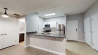 This single-floor unit offers a thoughtfully designed layout with a clean, contemporary aesthetic throughout. The kitchen is the centerpiece of the space, featuring white shaker-style cabinetry, a striking hexagonal tile backsplash, and a full suite of stainless steel appliances including a refrigerator, electric range, and built-in microwave. A peninsula counter with a dark countertop and integrated sink provides additional prep space and casual seating, seamlessly connecting the kitchen to the living area. Light wood-look flooring and recessed flush lighting lend a bright, welcoming feel to this central zone. The bedrooms are finished with plush wall-to-wall carpeting in a warm neutral tone, and each room benefits from natural light filtering through blinds-covered windows. One bedroom features a ceiling fan with a built-in light fixture, adding both comfort and function. The bathroom is well-appointed with a tub-shower combo, a modern vanity topped with a white countertop, a round mirror, and clean tile flooring laid in a herringbone pattern. Neutral gray walls are consistent throughout the unit, contributing to a cohesive, modern atmosphere that is both understated and inviting.