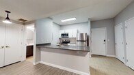 This single-floor unit offers a thoughtfully designed layout with a clean, contemporary aesthetic throughout. The kitchen is the centerpiece of the space, featuring white shaker-style cabinetry, a striking hexagonal tile backsplash, and a full suite of stainless steel appliances including a refrigerator, electric range, and built-in microwave. A peninsula counter with a dark countertop and integrated sink provides additional prep space and casual seating, seamlessly connecting the kitchen to the living area. Light wood-look flooring and recessed flush lighting lend a bright, welcoming feel to this central zone. The bedrooms are finished with plush wall-to-wall carpeting in a warm neutral tone, and each room benefits from natural light filtering through blinds-covered windows. One bedroom features a ceiling fan with a built-in light fixture, adding both comfort and function. The bathroom is well-appointed with a tub-shower combo, a modern vanity topped with a white countertop, a round mirror, and clean tile flooring laid in a herringbone pattern. Neutral gray walls are consistent throughout the unit, contributing to a cohesive, modern atmosphere that is both understated and inviting.