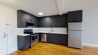 Located at 345 Main St in White Plains, NY, this single-floor residence offers a well-designed layout with modern finishes throughout. The kitchen is a standout feature, showcasing dark textured cabinetry that extends to the ceiling, crisp white countertops, and a full suite of stainless steel appliances including an electric range, over-the-range microwave, dishwasher, and refrigerator. Warm hardwood flooring flows seamlessly throughout the entire unit, creating a cohesive and inviting atmosphere. The open living area benefits from natural light streaming through large windows, which also provide views of the surrounding neighborhood. A hallway connects the living space to the bedroom, which features dual closets offering ample built-in storage, as well as direct access to additional rooms. The bathroom has been thoughtfully renovated with marble-style tile covering the floors and walls, a glass-enclosed walk-in shower, a wall-mounted vanity with a sleek vessel sink, and a mirrored medicine cabinet. Light gray walls throughout the unit complement the warm tones of the hardwood floors, resulting in a clean, contemporary aesthetic that is both functional and stylish.
