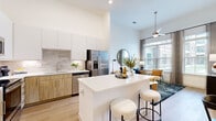 This thoughtfully designed one-bedroom, one-bathroom unit offers an efficient and stylish single-floor layout that maximizes every square foot. The open-concept kitchen and living area forms the heart of the space, anchored by a sleek kitchen featuring two-tone cabinetry—upper white cabinets contrasting warm wood-grain lower cabinets—paired with a neutral tile backsplash and stainless steel appliances. A white waterfall-edge island with bar seating provides both additional prep space and a natural transition into the living area.

The living room benefits from soaring ceilings and large, light-filled windows that frame outdoor views and flood the space with natural light. A modern ceiling fan adds both function and style overhead. Light-toned hardwood flooring runs throughout the main living areas, contributing to the bright, airy atmosphere.

The bedroom features plush carpeting, a generous window with outdoor views, and direct access to both the bathroom and a dedicated walk-in closet equipped with a full-width hanging rod and upper shelf storage. The bathroom includes light wood vanity cabinetry, a white countertop, a large mirror, and a tub-shower combination with decorative tile surround. Clean white walls and consistent warm-toned finishes unify the space throughout.