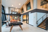 This thoughtfully designed amenity space blends industrial architecture with contemporary interiors across a single level. The dramatic main lobby greets visitors with soaring double-height ceilings, floor-to-ceiling glazing that floods the space with natural light, and exposed concrete columns that lend a raw, structural character. Polished concrete flooring runs throughout, complemented by warm wood accents on the staircase and mezzanine-level framing. Comfortable mid-century-inspired lounge chairs are arranged near the entrance, creating an inviting seating area with views to the outdoors.

Adjacent to the lobby, a spacious recreation room opens via wide sliding glass partitions, revealing a pool table, vibrant wallcoverings, and plush orange sofas. A foosball table and ping-pong table occupy the main common area, reinforcing the social, activity-driven character of the space.

The fitness center is a standout feature, finished with a sleek black ceiling adorned with striking yellow neon ring and linear lighting fixtures. A full row of Matrix treadmills lines the exterior glass wall, offering outdoor views during workouts. The free weights area is generously stocked with dumbbells, barbells, and benches, with floor-to-ceiling mirrors amplifying the space. Rubber flooring with teal directional markings adds both functionality and visual interest throughout the gym.