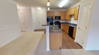 This single-floor residence features an open-concept layout that connects the kitchen and living area, creating a cohesive and functional living space. The kitchen is outfitted with honey-toned wood cabinetry offering generous storage, dark granite countertops, and a suite of black appliances including a refrigerator, range, microwave, and dishwasher. A peninsula island with matching granite and a decorative white column base extends into the space, providing additional prep and serving space with room for seating. Pendant lights hang above the peninsula, adding focused illumination and a subtle design detail. The kitchen features hardwood-style flooring, while the adjacent living area transitions to plush neutral carpeting, clearly delineating the two zones. Crown molding runs along the ceiling throughout, lending a polished, finished look to the interior. Multiple white panel doors lead to additional rooms and storage areas, visible from the main living space. The overall ambiance is clean and practical, with warm wood tones and neutral finishes creating a welcoming, move-in-ready environment.