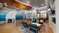 This single-floor amenity space showcases a cohesive coastal design theme carried throughout its open layout. The main game and lounge area is anchored by a full-size billiards table positioned beneath a striking curved wood-panel ceiling inset, complemented by modern ring chandelier lighting. A sculptural wave motif arches dramatically along one wall, reinforcing the surf-inspired aesthetic. The accent walls feature graduated blue shiplap paneling transitioning from white to deep teal, evoking the look of ocean waves. Decorative full-length surfboards flank a framed gallery wall, adding authentic beachside character.

The lounge seating area includes leather armchairs with gold-frame bases arranged around an oval ottoman, centered on a patterned area rug. A wall-mounted flat-screen television is positioned above a sleek linear fireplace, with circular metallic wall art above. Adjacent to the billiards area, a casual dining zone provides a round table with chairs, while bar-height seating lines a counter beneath the gallery wall. Large windows and a glass door offer views of an outdoor pool area, drawing in natural light. Wood-look tile flooring ties the entire space together, creating a warm, resort-style ambiance suited for social gatherings.