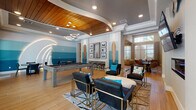 This single-floor amenity space showcases a cohesive coastal design theme carried throughout its open layout. The main game and lounge area is anchored by a full-size billiards table positioned beneath a striking curved wood-panel ceiling inset, complemented by modern ring chandelier lighting. A sculptural wave motif arches dramatically along one wall, reinforcing the surf-inspired aesthetic. The accent walls feature graduated blue shiplap paneling transitioning from white to deep teal, evoking the look of ocean waves. Decorative full-length surfboards flank a framed gallery wall, adding authentic beachside character.

The lounge seating area includes leather armchairs with gold-frame bases arranged around an oval ottoman, centered on a patterned area rug. A wall-mounted flat-screen television is positioned above a sleek linear fireplace, with circular metallic wall art above. Adjacent to the billiards area, a casual dining zone provides a round table with chairs, while bar-height seating lines a counter beneath the gallery wall. Large windows and a glass door offer views of an outdoor pool area, drawing in natural light. Wood-look tile flooring ties the entire space together, creating a warm, resort-style ambiance suited for social gatherings.
