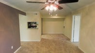 This single-floor space offers an open and functional layout centered around a spacious living area finished with wall-to-wall carpeting in a warm, neutral tone. The room is illuminated by a ceiling fan with an integrated light fixture, which serves as a practical focal point overhead. Crown molding runs along the upper perimeter of the living area, adding a subtle architectural detail that enhances the overall finish of the space.

A notable feature is the pass-through window connecting the living area to the kitchen, providing both visual openness and convenient access between the two spaces. The kitchen, visible through the pass-through, is equipped with black appliances including a microwave and stove, set against dark countertops. The kitchen area also features a raised counter ledge suitable for casual dining or serving.

The walls are finished in a soft, neutral beige tone that complements the carpeting and contributes to a cohesive, understated aesthetic throughout. HVAC vents are integrated into both the ceiling and wall, ensuring climate control across the space. A sliding door, partially visible on the right side, provides access to an adjacent area or storage space.