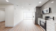 This contemporary single-floor space showcases a clean, modern design with an open-concept layout that seamlessly connects the living area and kitchen. Wood-look flooring flows throughout, complemented by crisp white walls and recessed lighting that creates a bright, welcoming ambiance. A ceiling fan and a large sliding glass door with horizontal shutter blinds allow natural light to fill the main living space, enhancing the airy feel.

The kitchen is a standout feature, equipped with stainless steel appliances including a Samsung range, over-the-range microwave, refrigerator, and dishwasher. High-gloss gray upper cabinetry contrasts elegantly with white lower cabinets, while a marble-style quartz backsplash and countertops add a refined, upscale touch. Ample cabinet storage extends to the ceiling, maximizing functionality.

The bathroom continues the marble aesthetic with floor-to-ceiling Calacatta-style tile walls, dark tile flooring, and a frameless sliding glass shower enclosure over a soaking tub, finished with a rain showerhead for a spa-like experience.

An additional room offers a versatile, light-filled space with hardwood-style flooring and access to the sliding door, ideal for use as a bedroom or flex room. The overall design presents a cohesive, modern aesthetic throughout.