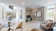 This well-appointed living room showcases a refined coastal-modern aesthetic through a thoughtful blend of architectural details and curated furnishings. Light-toned wood-look tile flooring runs throughout, lending a clean, airy feel to the entire space. A striking white shiplap accent wall serves as a central architectural feature, anchoring the room and providing a visual division between seating areas. Crown molding adds a polished finishing touch along the ceiling line.

Recessed lighting and a sculptural cluster pendant light fixture illuminate the space, complementing the natural light that filters in through tall, floor-to-ceiling windows dressed with sheer drapes. A built-in bench seat with a textured blue-gray upholstered backboard runs along one wall beneath a wall-mounted flat-screen television, flanked by woven rattan chairs and round marble-top accent tables — ideal for casual gathering.

A dedicated beverage station with a sage-green cabinet, under-counter refrigerator, and countertop appliances adds functional convenience. Abstract landscape and marble-swirl framed artwork is displayed throughout, contributing to an elevated, gallery-like ambiance. A separate seating nook features two modern accent chairs, a soft area rug, and additional curated wall art, offering a quieter retreat within the space.