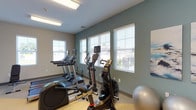 This fitness center is designed to support a comprehensive workout experience within a bright, functional layout. The space is organized to accommodate both cardio and strength training, with distinct areas dedicated to each discipline. The walls are painted in a combination of soft beige and muted teal-green tones, lending the room a calm, motivating atmosphere. Light-colored carpeted flooring runs throughout, providing comfort underfoot.

The cardio area is well-stocked with multiple treadmills, elliptical trainers, and an air resistance bike, all positioned along a windowed wall fitted with white horizontal blinds. Multiple windows allow generous natural light to fill the space while offering views of the surrounding exterior. Overhead, fluorescent light fixtures provide consistent illumination throughout.

The strength training section includes a multi-functional cable machine suitable for a wide range of resistance exercises, a dumbbell rack with a full set of free weights, and an adjustable weight bench. Additional accessories such as a stability ball are also present.

Two doorways along the far wall lead to what appear to be restroom or changing facilities, enhancing the practicality of the fitness center. The space is thoughtfully arranged to allow comfortable movement between equipment.