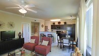 This single-floor residence showcases a thoughtfully designed open-concept layout that brings together the living, dining, and kitchen areas in one cohesive, flowing space. The living area is anchored by comfortable seating and a ceiling fan with light fixture, adding both function and a casual elegance to the room. Crown molding details along the upper walls lend a polished architectural touch throughout.

The kitchen is appointed with dark espresso cabinetry, a black refrigerator, electric range, and built-in microwave. A raised breakfast bar with seating provides a convenient casual dining option and serves as a natural divider between the kitchen and the main living area, while keeping the space open and connected.

Adjacent to the kitchen, a dedicated dining area features a dark-toned table and chair set, offering a more formal setting for meals. The combination of light-toned flooring and neutral walls allows natural light to move freely throughout the space, creating a bright and welcoming atmosphere. A hallway leads to additional rooms, suggesting a practical and well-organized floor plan. Overall, the space balances comfort and functionality with contemporary design elements, making it an inviting and versatile environment.