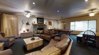 This single-floor space is designed with a warm, rustic aesthetic that creates an inviting atmosphere for both relaxation and entertainment. The central living area is anchored by a striking floor-to-ceiling stone fireplace, crafted from natural river rock, which serves as the room's defining architectural feature. A large, plush leather sectional sofa surrounds a wooden coffee table, providing ample seating for gatherings. Recessed lighting and a ceiling fan with an integrated light fixture illuminate the space efficiently, while decorative wall art — including nature-themed pieces and framed photography — reinforces the cabin-inspired character of the room. Adjacent to the seating area, a full-sized billiards table takes center stage, lit by a classic hanging pendant light fixture, making it a natural hub for recreational activity. A credenza along one wall offers additional storage and display space, accented by a small lamp and floral arrangement. Large windows allow natural light to filter in, brightening the darker tones of the carpet and ceiling. The overall ambiance is cozy yet spacious, blending rustic charm with functional design elements suited for leisure and social entertaining.