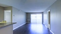 This single-floor space offers a clean and functional layout with a cohesive, understated design. The living area is the centerpiece of the unit, showcasing rich, dark hardwood flooring that reflects natural light and adds warmth to the room. The walls are finished in a neutral gray tone, complementing the overall modern and practical aesthetic.

A prominent sliding glass door, dressed with vertical blinds, serves as the focal point of the living area, inviting abundant natural light and providing access to the exterior. A wall-mounted air conditioning unit is positioned beneath the sliding door, ensuring year-round climate comfort. Baseboard heating is also present along the perimeter walls, offering an additional heating source.

The ceiling features a textured popcorn finish, a classic detail that adds dimension overhead. White baseboards and trim provide a crisp contrast against the gray walls and dark flooring. A partial countertop or bar surface is visible at the edge of the frame, hinting at a connected kitchen or dining area nearby.

The space is open and uncluttered, offering a versatile canvas for a variety of furniture arrangements. Overall, the unit presents a straightforward, well-lit living environment with durable finishes and practical built-in climate control features throughout.