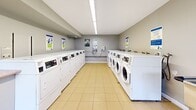 Located at 545-565 Belmont Ave W in Kitchener, Ontario, this shared laundry facility is a well-organized, single-floor space designed to efficiently serve building residents. The room is divided into two laundry sections, with commercial-grade front-loading washers and top-loading dryers arranged in neat rows along both sides of the facility. Instructional signage is posted throughout to guide users in operating the machines. The flooring throughout is finished in light beige ceramic tile, contributing to a clean and functional aesthetic. Walls are painted in a neutral grey tone, maintaining a bright and orderly environment. Overhead fluorescent lighting ensures the space is well-lit at all times. A stainless steel utility sink and a waste bin are conveniently positioned near the entrance, alongside a mounted fire extinguisher for safety. A notable feature is a windowed alcove at one end of the room, fitted with large multi-pane windows that frame views of the surrounding greenery and outdoor grounds, allowing natural light to fill the space. Baseboard heating runs beneath the windows, ensuring year-round comfort. The facility is accessed through a solid entry door and is thoughtfully equipped to meet the day-to-day laundry needs of the building's residents.