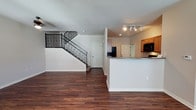 This two-story space offers an open-concept layout that connects the main living area and kitchen on the ground floor. Dark hardwood-style flooring runs throughout, providing a warm, grounding contrast to the crisp white walls and adding a sense of cohesion across the space.

The kitchen is positioned adjacent to the main living area and features warm-toned wood cabinetry, granite countertops, and black appliances including a built-in microwave. A peninsula counter extends into the open area, creating a natural divide between the kitchen and living space while maintaining the open feel. Track lighting above the kitchen provides focused illumination.

A standout architectural feature is the staircase, which rises from the main level to the second floor. It is framed by sleek black iron balusters and a clean white stringer, lending a modern, refined character to the interior. A door beneath the staircase offers additional utility or storage access.

The main living area is generously proportioned, offering flexibility in furniture arrangement. Recessed lighting supplements the overall brightness of the space, contributing to a clean, functional, and well-lit environment throughout both levels.
