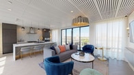 This elegantly designed single-floor space seamlessly integrates an open-concept living area and a chef's kitchen, creating a cohesive and functional layout bathed in natural light. The living area is anchored by a statement pendant light with a sculptural, ribbed shade in muted grey and gold, complementing the richly upholstered navy blue velvet accent chairs and a plush grey sofa accented with colorful throw pillows. A marble-topped coffee table and gold-accented side tables add refined detail, while a wall-mounted television flanked by brass sconces serves as a focal point.

The kitchen is a standout feature, centered around a generously proportioned waterfall island clad in striking white-veined stone. Dark espresso cabinetry contrasts beautifully with the bright stone backsplash, while a professional-grade range, stainless steel range hood, and double wall ovens provide serious culinary capability. A series of smoked glass pendant lights illuminates the island, where gold-framed upholstered barstools complete the refined aesthetic.

Floor-to-ceiling black-framed glass doors open onto a private balcony, flooding the interior with daylight and offering sweeping elevated views of the surrounding cityscape. Light herringbone-patterned tile flooring and a slatted white ceiling add architectural texture throughout, lending the space a polished, contemporary character.