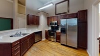 This single-floor unit offers a thoughtfully laid-out interior defined by warm hardwood flooring that runs throughout every room, creating a cohesive and inviting atmosphere. The entryway, framed by a distinctive multi-pane dark wood door with transom detail, leads into a central hallway that connects the main living areas. The kitchen is a functional highlight of the space, featuring rich dark wood cabinetry, light-toned countertops, and a full suite of stainless steel appliances including a gas range, built-in microwave, dishwasher, and a large French door refrigerator. A peninsula-style breakfast bar separates the kitchen from the adjoining area, and a suspended linear pendant light adds a modern touch beneath the dark painted ceiling with crown molding. The living and flex rooms showcase dark wood wainscoting along the lower walls, crown molding at the ceiling, and large windows fitted with horizontal blinds that allow natural light to filter in. A decorative globe pendant light fixture anchors each room. The bathroom is clean and well-appointed, with a full tub and shower combination featuring marble-style surround walls, grab bars, and hardwood flooring continuing into the space. Overall, the unit blends classic architectural character with functional modern updates.
