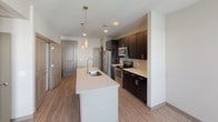 This single-floor unit offers a thoughtfully designed layout that balances style and functionality throughout. The kitchen is a standout feature, showcasing rich dark cabinetry that extends to the ceiling, light quartz countertops, and a full suite of stainless steel appliances including a French door refrigerator with ice maker, electric range, and built-in microwave. A large central island with an integrated sink and pendant lighting above serves as both a prep area and a casual gathering spot. Wood-look flooring runs through the main living and kitchen areas, adding warmth and visual continuity. A dedicated pantry closet adjacent to the kitchen provides generous additional storage. The bedroom is finished with plush carpeting and features large windows dressed with blinds, allowing for ample natural light and views of the surrounding exterior. A ceiling fan with a light fixture adds comfort to the space. The bathroom is clean and practical, featuring a walk-in shower with glass enclosure, a vanity with cabinetry, wood-look flooring, and grab bar accents that speak to accessible design. Wire shelving in the closet area offers well-organized storage. Overall, the unit presents a contemporary aesthetic with neutral tones and modern finishes throughout.