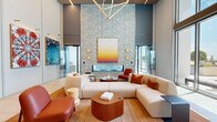 Located at 1000 Spring St NW in Atlanta's Midtown neighborhood, this expansive amenity space spans a single floor and offers a thoughtfully designed environment built for both relaxation and entertainment. The centerpiece is a dramatic double-height lobby lounge distinguished by soaring ceilings, floor-to-ceiling glazing, and an abundance of natural light that floods the interior. A bold botanical-patterned accent wall frames a sleek linear fireplace, while a statement geometric chandelier in brushed gold anchors the space above a curated seating arrangement of curved cream sectionals and rich terracotta leather chairs. Large-format porcelain tile flooring transitions seamlessly between zones, complemented by warm wood accents throughout. Curated artwork lines the walls, adding a gallery-like quality to the corridors and common areas. The game room is a standout feature, outfitted with a billiards table, foosball table, shuffleboard, and comfortable lounge seating arranged atop a vibrant geometric-patterned carpet, all bathed in layered pendant lighting. A dedicated indoor golf simulator room provides an immersive experience with a full projection screen and putting turf. A more intimate lounge area with a deep navy sofa and warm accent lighting connects seamlessly to the game room, offering a versatile retreat within this well-appointed amenity floor.