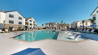 Located at 1001 Victory Lap Ln in Holly Ridge, North Carolina, this outdoor amenity area showcases a generously sized resort-style swimming pool as its centerpiece. The pool features a smooth, light-toned concrete surround with easy-entry staircases and is enclosed by a sleek black iron fence, ensuring both safety and an open, airy feel. Surrounding the pool, an expansive deck accommodates numerous white lounge chairs and blue-umbrella shaded seating areas, providing ample space for sunbathing and relaxation. Lush palm trees are strategically planted throughout the deck, lending a coastal, vacation-inspired ambiance that complements the sunny North Carolina setting. A covered clubhouse pavilion anchors one side of the pool area, featuring board-and-batten white siding, stone accents, and large windows that bring in abundant natural light. The pavilion includes an outdoor dining area with tables and chairs, ideal for social gatherings. Manicured turf patches accent the deck, adding pops of greenery between lounge areas. The multi-story residential buildings surrounding the pool feature contemporary coastal architecture with neutral siding, balconies, and large windows. The entire outdoor space is designed to evoke a relaxed, community-centered lifestyle under the open Carolina sky.