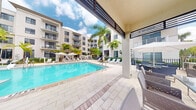 This outdoor lounge is a well-appointed open-air amenity space designed for leisure and relaxation. At its centerpiece is a large, crystal-clear swimming pool framed by a broad, light-toned paver deck that extends generously around the water's edge. The pool area is lined with lounge chairs and white canopy umbrellas, providing ample shaded spots for sunbathing and unwinding. Tall, mature palm trees are strategically placed throughout the courtyard, lending a distinctly tropical ambiance to the setting. A covered pergola-style structure along one side of the lounge offers a partially shaded dining and seating zone, furnished with contemporary weather-resistant chairs and tables — ideal for outdoor gatherings. The architectural backdrop consists of a multi-story residential building with balconies overlooking the pool courtyard, creating an enclosed, private atmosphere. The open sky above floods the space with abundant natural light, enhancing the bright and airy feel. The combination of lush greenery, modern furnishings, and elegant hardscaping makes this outdoor lounge a sophisticated and inviting communal space suited for both quiet relaxation and social interaction.