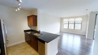 This single-floor residence features one bedroom and one and a half bathrooms, offering a functional and modern living environment. The open-concept layout connects the kitchen and living area seamlessly, creating a cohesive and spacious feel throughout.

The kitchen is outfitted with rich, dark-stained wood cabinetry that provides ample storage, complemented by sleek dark granite countertops that extend to a peninsula with an integrated stainless steel sink. The peninsula serves as a natural divider between the kitchen and the adjoining living space, offering additional prep area and casual seating potential. A dishwasher is integrated beneath the counter, contributing to the kitchen's streamlined aesthetic. Tile flooring anchors the kitchen zone.

The living area transitions to warm hardwood-style flooring, lending a refined and welcoming ambiance. A large window along the far wall allows natural light to fill the space, offering views of the surrounding greenery. Neutral wall tones throughout contribute to a bright, clean atmosphere. The overall design balances practicality with a polished, contemporary aesthetic.