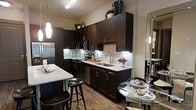 This single-floor space showcases a thoughtfully designed kitchen as its centerpiece. Rich, dark espresso cabinetry extends from floor to ceiling, providing generous storage while creating a bold visual contrast against the light-toned quartz countertops. A classic subway tile backsplash with a subtle linear pattern adds texture and dimension to the cooking area, complemented by under-cabinet lighting that illuminates the workspace effectively. Stainless steel appliances, including a refrigerator, oven, and dishwasher, lend a professional quality to the kitchen. A kitchen island with a white countertop and built-in seating via black bar stools offers additional prep space and casual dining. Track lighting overhead ensures the kitchen remains bright and functional. Hardwood-style flooring flows throughout, adding warmth and cohesion to the space. Adjacent to the kitchen, a compact dining area accommodates a glass-top table with seating, making efficient use of the available space. The overall design balances dark, dramatic cabinetry with lighter surfaces, creating a contemporary and inviting atmosphere suitable for both cooking and entertaining.