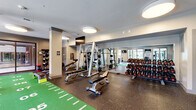 Located at 1015 Nelson Merry Street in Nashville, TN, this generously sized fitness center spans a single floor and is thoughtfully divided into multiple distinct workout zones, offering a comprehensive exercise experience. The cardio area features a row of treadmills, elliptical machines, and stationary bikes positioned along large windows that flood the space with natural light and provide views of surrounding greenery. A dedicated strength training zone is outfitted with a full array of Precor selectorized machines, free weights, and cable systems, all set against mirrored walls that enhance the sense of openness. At the heart of the facility lies a standout functional training area showcasing a turf sprint track marked with yardage lines, ideal for agility and conditioning drills. An adjoining studio space features cork flooring, textured accent walls, a wall-mounted display, and a glass garage door that opens to an outdoor deck, creating a bright, flexible environment for group classes or personal training. Ceiling-mounted circular flush lighting throughout gives the facility a clean, modern aesthetic. The durable rubber flooring in primary workout areas, combined with the variety of specialized zones, makes this fitness center a well-equipped destination for all training styles.