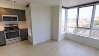 This single-floor space showcases a clean, modern design with an open-concept layout that maximizes both functionality and natural light. The living area features light wood laminate flooring that runs throughout, lending warmth to the otherwise minimalist aesthetic defined by crisp white walls and simple white baseboards.

A standout feature is the large corner window arrangement, which floods the living space with natural light and offers expansive views of the surrounding urban landscape. The elevated positioning of the unit enhances these views, providing an open, airy feel.

A partial room divider wall creates a subtle separation between the living area and the kitchen zone, adding architectural interest without fully closing off the space. The kitchen area is visible to one side and features dark cabinetry with sleek hardware, providing a striking contrast against the lighter tones of the living area.

The layout is thoughtfully efficient, making strong use of available space. The combination of natural light, contemporary finishes, and a neutral color palette creates a versatile, move-in ready environment well-suited to a variety of interior styles.