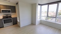 This single-floor space showcases a clean, modern design with an open-concept layout that maximizes both functionality and natural light. The living area features light wood laminate flooring that runs throughout, lending warmth to the otherwise minimalist aesthetic defined by crisp white walls and simple white baseboards.

A standout feature is the large corner window arrangement, which floods the living space with natural light and offers expansive views of the surrounding urban landscape. The elevated positioning of the unit enhances these views, providing an open, airy feel.

A partial room divider wall creates a subtle separation between the living area and the kitchen zone, adding architectural interest without fully closing off the space. The kitchen area is visible to one side and features dark cabinetry with sleek hardware, providing a striking contrast against the lighter tones of the living area.

The layout is thoughtfully efficient, making strong use of available space. The combination of natural light, contemporary finishes, and a neutral color palette creates a versatile, move-in ready environment well-suited to a variety of interior styles.