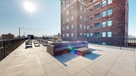 This generously scaled rooftop terrace offers an expansive outdoor amenity space positioned atop a multi-story brick building in an urban setting. The terrace is finished with large-format concrete pavers throughout, providing a clean, contemporary surface that extends across the full footprint of the roof. A sleek black metal perimeter railing frames the space while preserving unobstructed, panoramic views of the surrounding cityscape, including recognizable bridges and landmark architectural structures on the horizon.

The terrace is thoughtfully organized into multiple distinct zones. Several seating clusters featuring outdoor lounge furniture, including cushioned sofas, chairs, and low tables, are distributed across the space to accommodate both intimate gatherings and larger groups. A section of artificial turf lawn introduces a softened green element, offering a contrast to the hard-paved surfaces. String lights are strung overhead across pole mounts, providing ambient illumination for evening use.

A wood-clad mechanical enclosure with a rooftop access door anchors one end of the terrace, while the open sky above ensures abundant natural light throughout the day. The overall atmosphere is modern, social, and well-suited for outdoor entertaining with commanding urban views.
