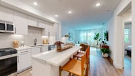 Located in Carlsbad, CA, this thoughtfully designed 2-bedroom, 3-bathroom single-floor residence offers a bright and contemporary living experience. The open-concept kitchen and living area serves as the heart of the home, where white shaker cabinetry with matte black hardware, a neutral tile backsplash, quartz countertops, and stainless steel appliances create a polished, modern aesthetic. A central island with bar seating flows seamlessly into the living room, which is bathed in natural light from large windows overlooking lush outdoor surroundings and warm wood-look flooring throughout ties the spaces together cohesively. The primary bedroom is a generous retreat featuring a serene accent wall, ceiling fan, and a spacious walk-in closet with multi-tier shelving and hardwood flooring. The second bedroom is equally inviting, with large windows offering verdant outdoor views and abundant natural light. All three bathrooms are finished with clean white vanities, quartz surfaces, and matte black fixtures; the primary bath includes a glass-enclosed walk-in shower with tile surround. Additional highlights include a dedicated laundry area and ample closet storage, making this well-appointed residence as functional as it is stylish.