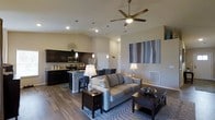This single-floor residence showcases an open-concept layout that effortlessly connects the kitchen, dining, and living areas into one cohesive, flowing space. Vaulted ceilings amplify the sense of volume and airiness throughout, while a ceiling fan with integrated lighting adds both comfort and a contemporary touch overhead.

The kitchen is outfitted with dark espresso cabinetry, stainless steel appliances, and a breakfast bar with seating, making it equally suited for casual dining and meal preparation. Pendant lighting above the bar adds a warm, inviting glow that complements the recessed lighting found throughout the space.

The living area is anchored by a neutral-toned sofa and coordinated dark wood furniture, creating an elegant yet comfortable atmosphere. Wood-look flooring runs throughout, providing a durable and visually appealing foundation that ties the entire open floor plan together. Neutral wall tones serve as a versatile backdrop, accented by decorative artwork that adds a splash of color and personality.

The layout promotes a natural flow between functional zones, making the space ideal for both everyday living and entertaining. Overall, the interior strikes a balance between modern design sensibilities and warm, welcoming ambiance.