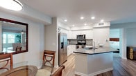 This thoughtfully designed single-floor residence offers a cohesive layout that balances open communal areas with comfortable private spaces. The heart of the home is a modern, open-concept kitchen outfitted with white shaker cabinetry, gray tile backsplash, stainless steel appliances including a French door refrigerator with ice dispenser, an electric range, and an over-the-range microwave. A generously sized island with dark-edged gray countertops provides ample workspace and a built-in sink, flowing seamlessly into the living area. The living room features light wood-style flooring, a ceiling fan, and direct sightlines to the kitchen, creating an inviting, connected atmosphere. A dedicated dining area with glass-top furniture adds further functionality to the shared living space. The bedrooms are carpeted and well-sized, each featuring ceiling fans and windows with natural light. The bathrooms are finished with white cabinetry, dark countertops, framed mirrors, and wood-style flooring. A separate office or flex room offers wood flooring and bright window exposure. An in-unit laundry closet houses a full-size side-by-side washer and dryer. Neutral tones and recessed lighting throughout lend the space a clean, modern character.
