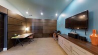 This single-floor space serves as a well-appointed business center designed to support productivity and focused work. The room makes a strong visual impression with its rich, dark wood wall paneling that spans the length of the space, providing a warm and professional backdrop. Contrasting the wood tones, a vibrant teal accent wall adds a modern, energizing touch to the environment.

The space features multiple dedicated workstations, each equipped with ergonomic task chairs, offering ample room for individual work. A built-in credenza with white cabinetry and a dark wood countertop runs along one wall, providing generous storage and surface space. This area also houses a wall-mounted flat-screen display, making it suitable for presentations or multimedia use.

Recessed lighting throughout the ceiling ensures the space is evenly and efficiently illuminated, complementing the clean, contemporary design aesthetic. Light-toned flooring ties the room together, balancing the darker wall elements. Decorative geometric wall art on the teal accent wall adds a subtle artistic element without distracting from the workspace's functional purpose. Overall, this business center combines practical amenities with a polished, modern design to create an environment conducive to both individual and collaborative work.
