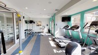 This modern fitness center is thoughtfully designed to support a comprehensive workout experience across several distinct zones. The space is anchored by a bright, open cardio area outfitted with a row of treadmills, elliptical machines, and a stair climber, all positioned to take advantage of natural light streaming through large windows. Bold teal accent walls provide a vibrant contrast to the crisp white ceilings and neutral-toned striped carpet flooring, creating an energizing atmosphere throughout.

A dedicated free weights area features a full dumbbell rack, a weight bench, and wall-mounted mirrors that enhance the sense of openness. Multiple flat-screen TVs are mounted throughout the space, keeping members entertained during their workouts. A cable and functional training machine anchors a central strength training section, offering versatility for a range of exercises.

The fitness center also includes a glass-partitioned studio room with light wood flooring, ideal for stretching, yoga, or cycling. This studio houses a spin bike, a rack of kettle bells, additional dumbbells, and a mounted display screen. The floor-to-ceiling glass walls with frosted stripe detailing allow visibility while maintaining a sense of separation. Recessed lighting throughout ensures consistent illumination, and the overall layout promotes smooth traffic flow across the entire facility.