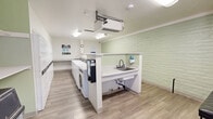 Located at 6300 Montgomery Blvd NE in Albuquerque, New Mexico, this shared laundry facility offers a clean, functional, and efficiently organized space designed for ease of use. The room features light wood-look vinyl flooring throughout and soft sage-green painted brick walls that lend a fresh, welcoming ambiance. Bright fluorescent overhead lighting ensures the space is well-lit at all times.

The main area is lined with a row of commercial-grade top-load washing machines paired with a corresponding bank of dryers stacked above, providing ample laundry capacity. A built-in folding counter runs along the perimeter, offering generous surface area for sorting and folding. A deep utility sink with a soap dispenser is conveniently positioned for pre-treating garments, accompanied by a mounted paper towel dispenser nearby.

A cushioned bench provides a comfortable spot for waiting, while a bulletin board on the wall displays informational notices and laundry instructions. A waste bin is centrally accessible for added convenience. Natural light filters in through a set of windows, complementing the bright interior. A wall-mounted HVAC unit helps maintain a comfortable temperature year-round. The space is thoughtfully laid out to accommodate multiple users simultaneously, making it a practical and well-maintained communal amenity.