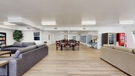 Located at 1560 Yosemite Dr in the Eagle Rock neighborhood of Los Angeles, this single-story community facility offers a generous and well-organized layout across one expansive floor. The interior is finished with light wood-look flooring throughout, crisp white walls, and bright fluorescent lighting that creates a clean, welcoming atmosphere.

The living area is divided into two distinct seating zones. One section features plush gray sofas arranged around a flat-screen television flanked by bookshelves, providing a comfortable space for relaxation and entertainment. The adjacent lounge area offers neutral-toned sofas and accent chairs positioned near large windows that invite abundant natural light and views of the surrounding greenery.

A communal dining area accommodates multiple dining tables and dark wood chairs, making it well-suited for group gatherings and meals. The mailroom area includes rows of individual resident mailboxes and additional dining seating, anchored by a central column decorated with hanging floral accents.

The dedicated laundry room is equipped with multiple washing machines and dryers, a folding counter, and a clean, utilitarian layout. Two ADA-compliant bathrooms are outfitted with grab bars, comfort-height fixtures, and light wood-look flooring, ensuring accessibility for all users. The overall design prioritizes functionality, comfort, and community engagement.