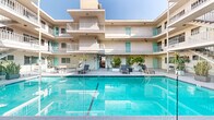 This multi-story courtyard property showcases a classic mid-century architectural style, characterized by clean stucco exterior walls, tiered balconies with white metal railings, and a cohesive color palette of warm beige and vibrant teal accent doors. The centerpiece of the property is a generously sized outdoor swimming pool, framed by glass panel barriers and surrounded by a wide, open pool deck. The deck is furnished with modern lounge chairs and planters featuring tropical palms and lush greenery, creating a resort-like ambiance bathed in abundant natural sunlight.

The building wraps in a U-shaped configuration around the courtyard, with multiple levels of private balconies overlooking the pool area. Ground-floor access corridors connect the surrounding units to the shared outdoor spaces, with decorative potted tropical plants lending a lively, welcoming character to the passageways. A covered interior corridor, accented with a chandelier and colorful tile artwork, provides a stylish transition between the pool courtyard and additional areas of the property.

Additional secondary courtyard spaces are also present, offering shaded, open-air areas with a more intimate feel. Throughout the property, the interplay of open sky, natural light, and consistent architectural detailing creates a cohesive and inviting communal environment.