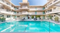 This multi-story courtyard property showcases a classic mid-century architectural style, characterized by clean stucco exterior walls, tiered balconies with white metal railings, and a cohesive color palette of warm beige and vibrant teal accent doors. The centerpiece of the property is a generously sized outdoor swimming pool, framed by glass panel barriers and surrounded by a wide, open pool deck. The deck is furnished with modern lounge chairs and planters featuring tropical palms and lush greenery, creating a resort-like ambiance bathed in abundant natural sunlight.

The building wraps in a U-shaped configuration around the courtyard, with multiple levels of private balconies overlooking the pool area. Ground-floor access corridors connect the surrounding units to the shared outdoor spaces, with decorative potted tropical plants lending a lively, welcoming character to the passageways. A covered interior corridor, accented with a chandelier and colorful tile artwork, provides a stylish transition between the pool courtyard and additional areas of the property.

Additional secondary courtyard spaces are also present, offering shaded, open-air areas with a more intimate feel. Throughout the property, the interplay of open sky, natural light, and consistent architectural detailing creates a cohesive and inviting communal environment.