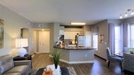 This single-floor residence showcases an open-concept layout that seamlessly connects the living area and kitchen, creating a spacious and functional environment. The kitchen is a standout feature, equipped with white upper cabinetry, dark countertops, and a peninsula that doubles as a breakfast bar, providing additional seating and prep space. Stainless steel and standard appliances, including a refrigerator, are neatly integrated into the design. A tile backsplash adds a subtle decorative element behind the cooking area. The flooring throughout is a rich hardwood-style material that adds warmth and continuity across the open space. Neutral gray walls provide a versatile backdrop that complements both the kitchen and living areas. A window with horizontal blinds allows natural light to filter into the space, brightening the interior. The entry area, visible from the main living space, features a classic paneled door flanked by warm overhead lighting. Recessed ventilation is placed thoughtfully throughout the ceiling, contributing to the clean, uniform aesthetic. Overall, the space balances modern design with practical, everyday functionality.