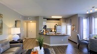 This single-floor residence showcases an open-concept layout that seamlessly connects the living area and kitchen, creating a spacious and functional environment. The kitchen is a standout feature, equipped with white upper cabinetry, dark countertops, and a peninsula that doubles as a breakfast bar, providing additional seating and prep space. Stainless steel and standard appliances, including a refrigerator, are neatly integrated into the design. A tile backsplash adds a subtle decorative element behind the cooking area. The flooring throughout is a rich hardwood-style material that adds warmth and continuity across the open space. Neutral gray walls provide a versatile backdrop that complements both the kitchen and living areas. A window with horizontal blinds allows natural light to filter into the space, brightening the interior. The entry area, visible from the main living space, features a classic paneled door flanked by warm overhead lighting. Recessed ventilation is placed thoughtfully throughout the ceiling, contributing to the clean, uniform aesthetic. Overall, the space balances modern design with practical, everyday functionality.